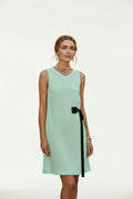 A woman wearing a sleeveless mint green dress with a black side bow, standing outdoors near a blue shutter and sea background while holding a woven straw bag.