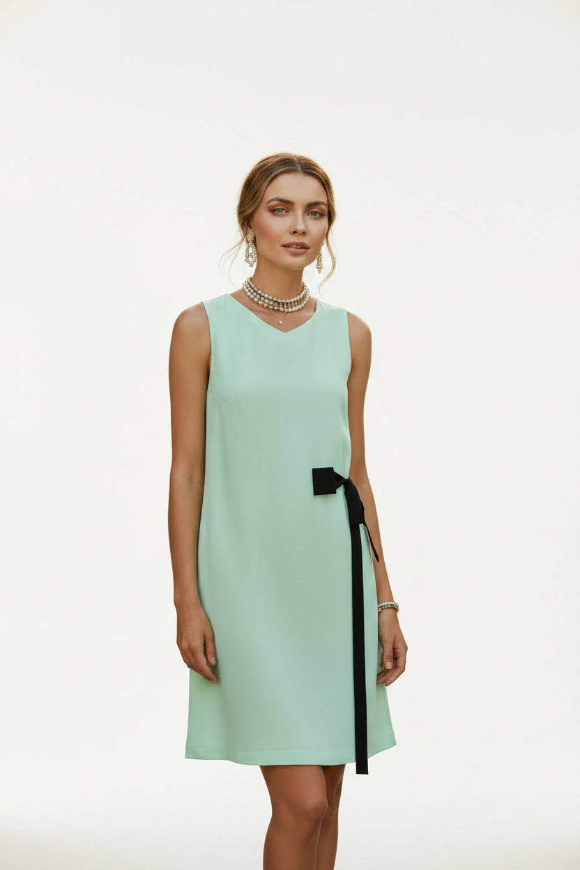 A woman wearing a sleeveless mint green dress with a black side bow, standing outdoors near a blue shutter and sea background while holding a woven straw bag.