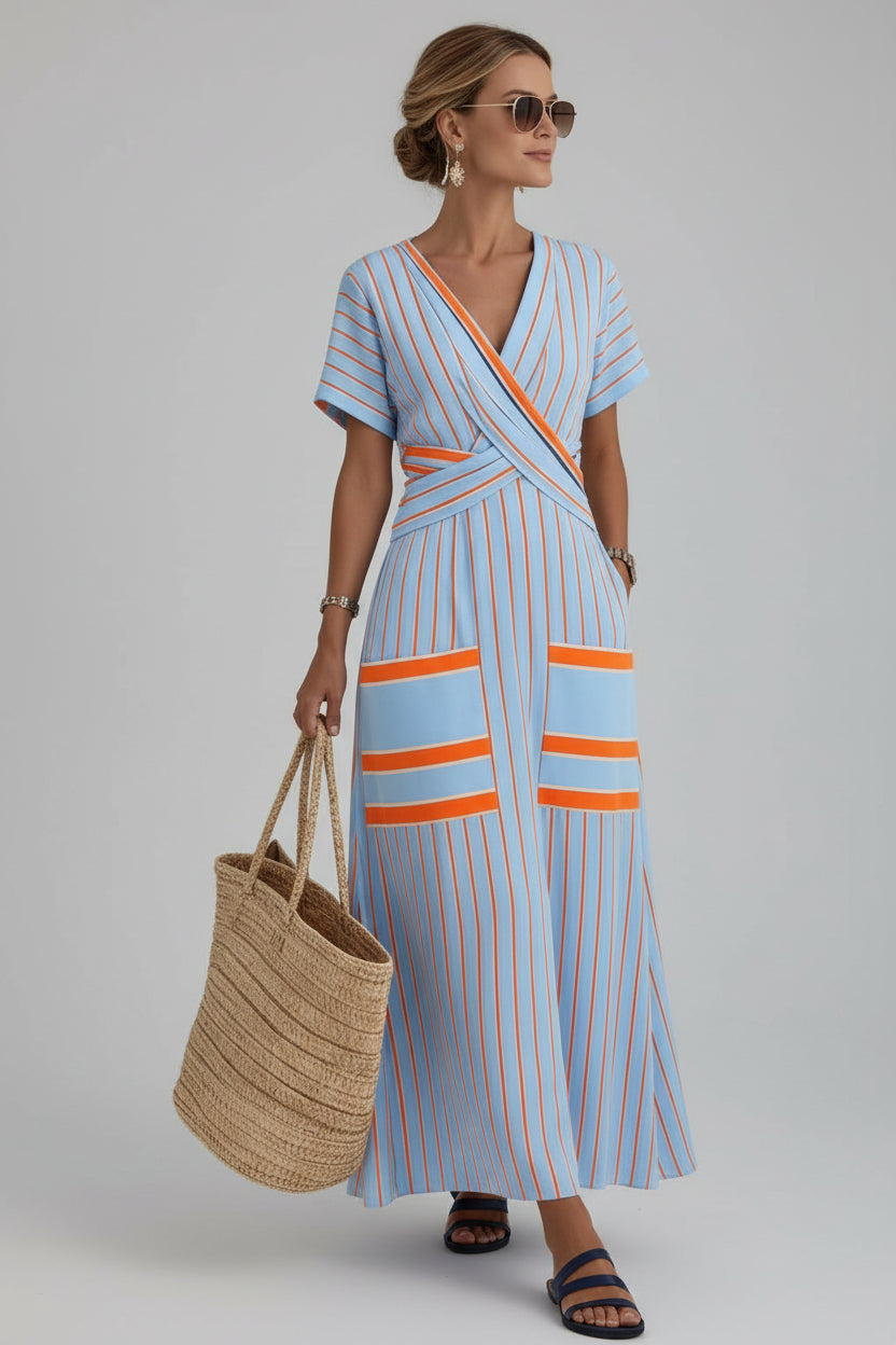 A woman wearing a light blue wrap-style maxi dress with orange and white vertical stripes walks along a sunlit white-walled street by the sea. She is carrying a large woven straw tote bag and wearing blue slide sandals.