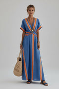 A woman wearing a blue and orange striped maxi dress walks along a stone path by the sea, holding a large woven straw tote bag. The background shows white buildings and the blue water.