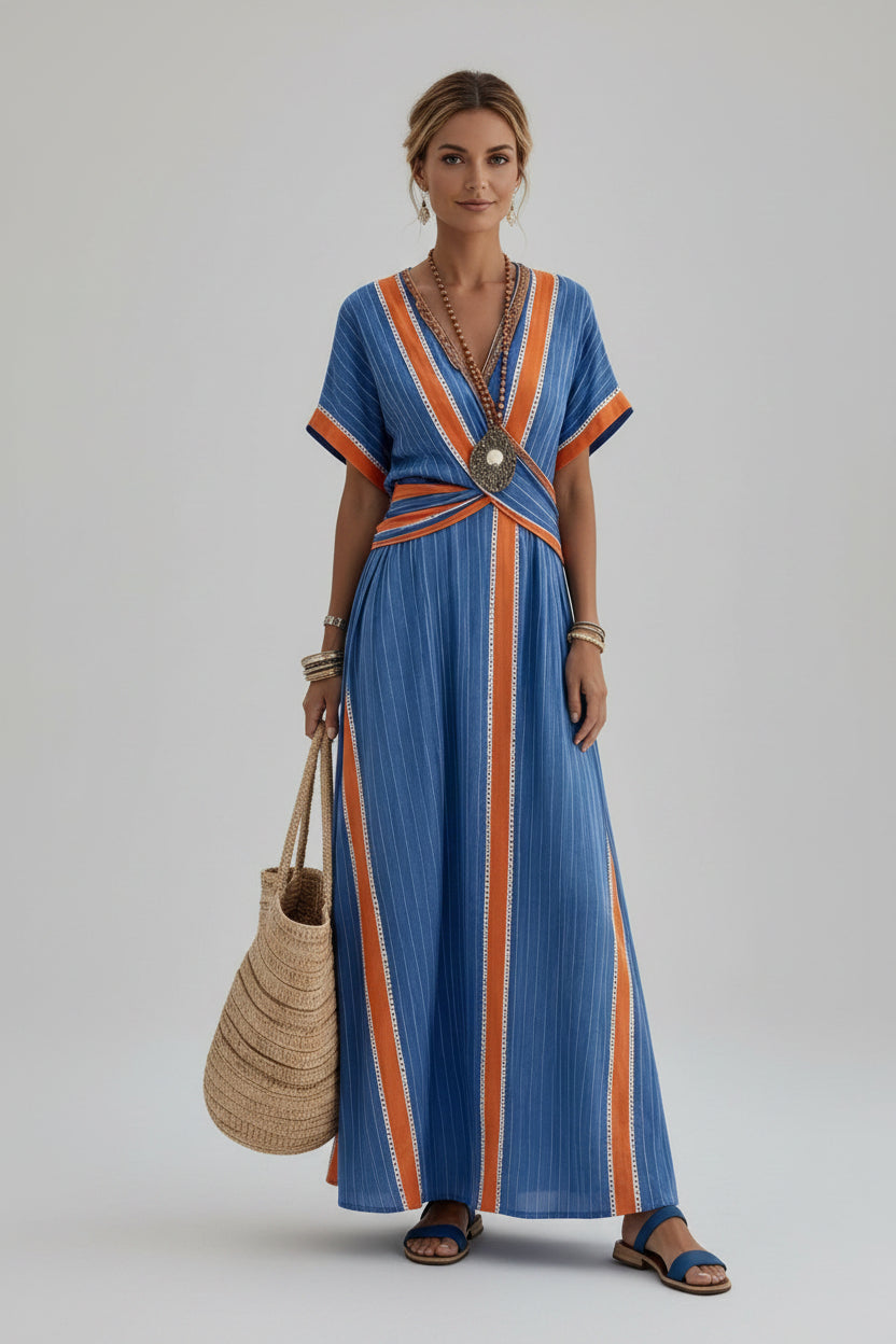 A woman wearing a blue and orange striped maxi dress walks along a stone path by the sea, holding a large woven straw tote bag. The background shows white buildings and the blue water.
