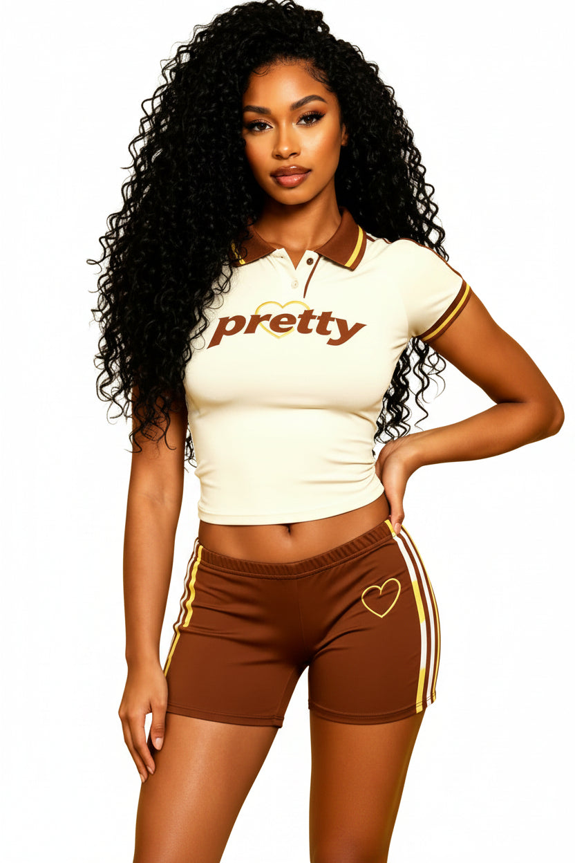 A woman modeling a fitted short-sleeve polo shirt and matching shorts set against a solid yellow background. The polo is cream with brown and gold trim and the word 'pretty' across the chest, while the matching brown shorts have gold and white side stripes and a gold heart graphic.