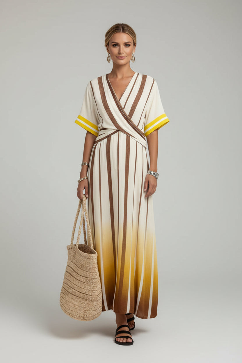 A woman wearing a wrap-style maxi dress with vertical brown and cream stripes and a yellow-to-cream gradient hem, walking down a white-walled street in a Mediterranean-style setting while carrying a woven straw tote bag.
