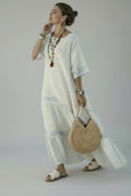 A woman wearing a white bohemian maxi dress with light blue floral patterns and tiered ruffles, standing on a white-walled coastal path with the sea and buildings in the background. She is holding a round woven straw handbag and wearing white slide sandals.