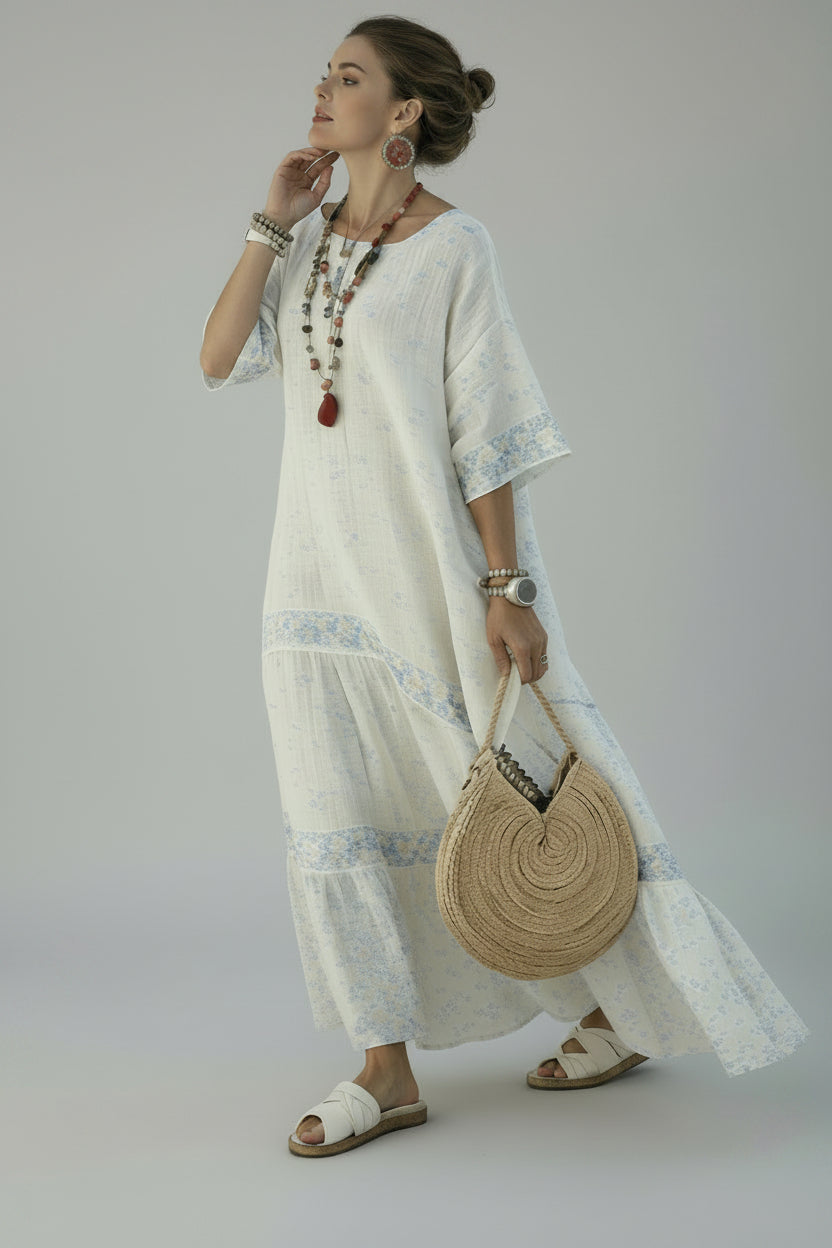 A woman wearing a white bohemian maxi dress with light blue floral patterns and tiered ruffles, standing on a white-walled coastal path with the sea and buildings in the background. She is holding a round woven straw handbag and wearing white slide sandals.