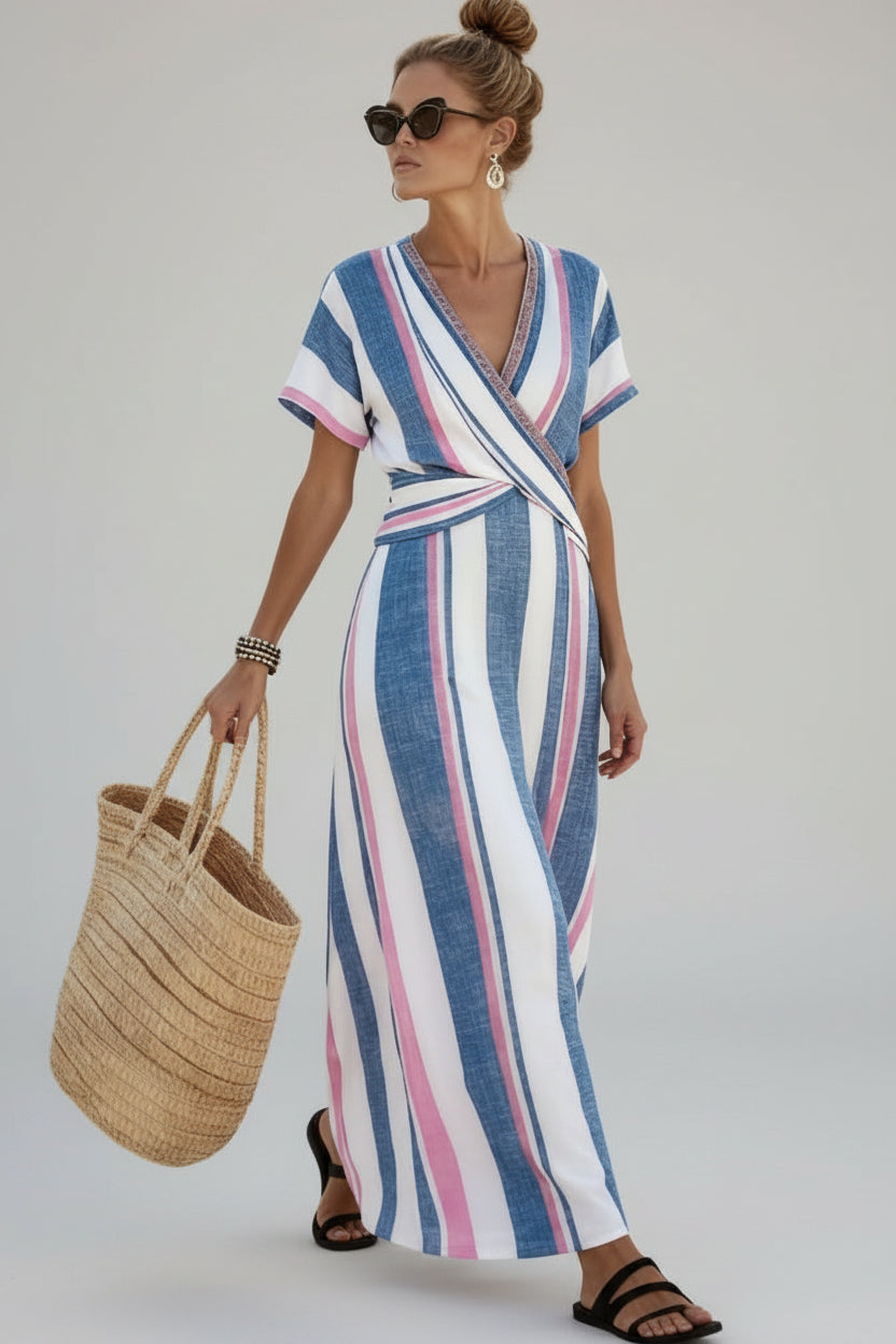 A woman wearing a blue, white, and pink vertically striped wrap maxi dress walks on a sandy beach path, holding a large woven straw tote bag.