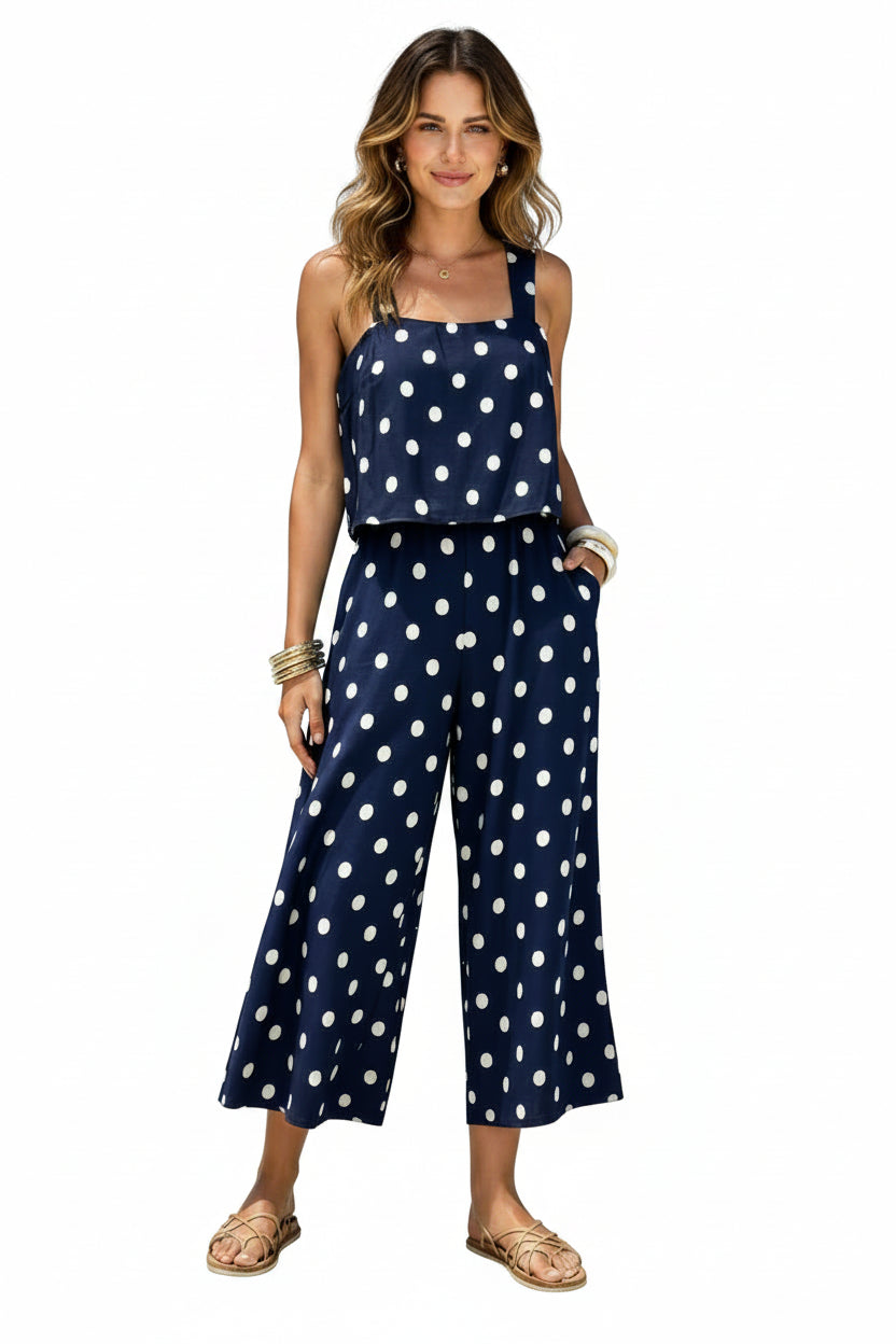 A woman wearing a navy blue and white polka dot sleeveless jumpsuit stands on a sandy beach with turquoise ocean water in the background.