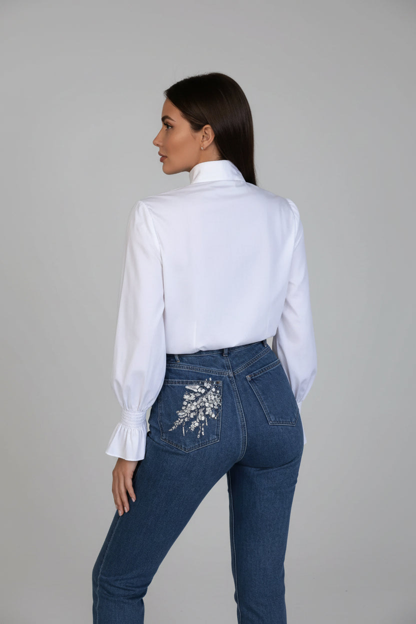 Person wearing a white shirt and blue jeans with floral embroidery in a room.